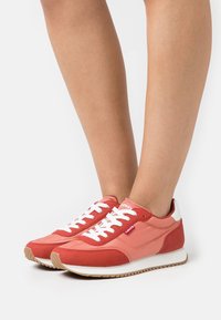 Baskets rouges et roses avec des lacets blancs et un logo, présentant un upper en tissu texturé et une semelle en caoutchouc, montrées sur une jambe nue.