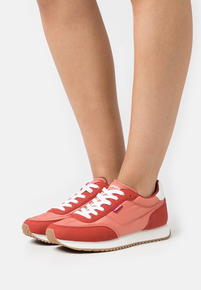Baskets rouges et roses avec des lacets blancs et un logo, présentant un upper en tissu texturé et une semelle en caoutchouc, montrées sur une jambe nue.