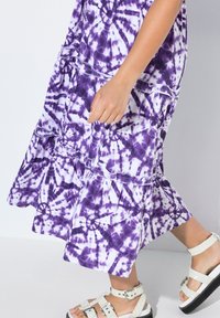 Lila und weiße Batik-Tunika mit einem gestuften Design, das mit Rüschenkanten versehen ist. Getragen mit weißen Sandalen, die verstellbare Riemen und eine schwarze Sohle haben.