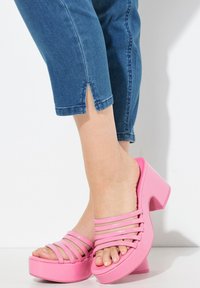 Vaaleanpunaiset korot, joissa on useita vaakasuoria hihnoja. Sileä, kiiltävä materiaali ja paksu korko. Yhdistetty vaaleisiin, lyhyisiin farkkuihin, joissa on sivuaukot.