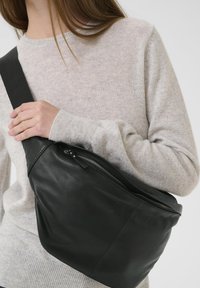 Borsa a tracolla in pelle nera con una texture liscia, dotata di chiusura con cerniera e una larga tracolla regolabile. Indossata sopra un maglione grigio chiaro.