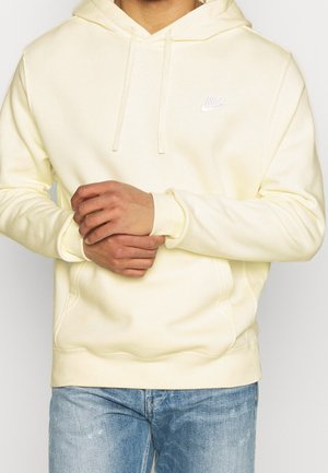 Homme portant un sweat à capuche Nike jaune clair avec cordons, tenant un poignet, assorti à un jean bleu clair.