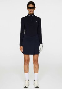 Granatowa koszulka polo z długim rękawem, białym kołnierzykiem i plisą na guziki, zestawiona z dopasowaną granatową spódnicą z przednim rozporkiem, noszona z rękawiczką i okularami przeciwsłonecznymi.