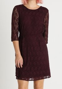 Robe en dentelle bourgogne avec des manches trois-quarts, motif floral et taille ajustée. La couche supérieure présente un revêtement en dentelle texturée.