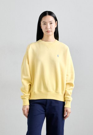 Sudadera de algodón amarilla con cuello redondo y puños de canalé, que presenta un pequeño logo azul en el pecho. Combinada con pantalones navy.