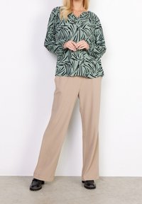 Blusa verde con motivo astratto nero, scollatura a V e maniche arricciate; abbinata a pantaloni beige a gamba larga e scarpe nere con suola spessa.
