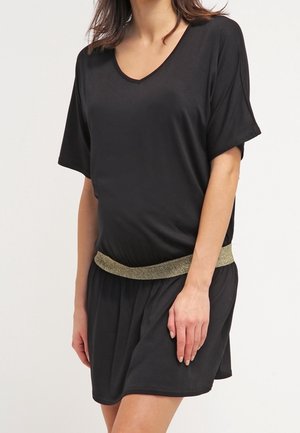 Vrouw in een zwarte jurk met korte mouwen en een gouden glinsterende tailleband, staand met haar rechterhand rustend op haar dij.