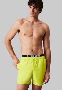 Neongele zwembroek gemaakt van lichtgewicht stof met een zwarte elastische tailleband waarop het Calvin Klein-logo staat, en een trekkoorddetail.