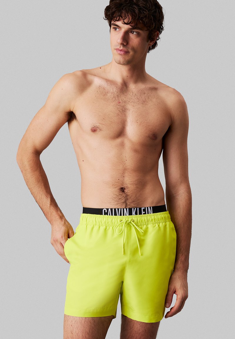Neongele zwembroek gemaakt van lichtgewicht stof met een zwarte elastische tailleband waarop het Calvin Klein-logo staat, en een trekkoorddetail.