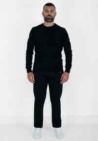 Maglione nero a maglia con scollo rotondo, abbinato a pantaloni neri. Il modello indossa sneaker bianche. Texture liscia, design aderente in tutta la lunghezza.