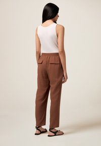 Débardeur blanc, pantalon marron taille élastique avec deux poches arrière, tissu texturé, et sandales noires avec des accents décoratifs.