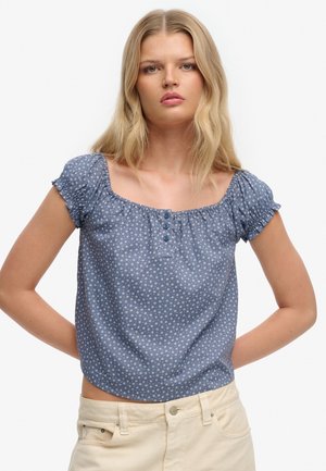 Lyshåret kvinde iført en blå blomsterprint kortærmet bluse med knapper og cremefarvede bukser, stående med hænderne bag ryggen.
