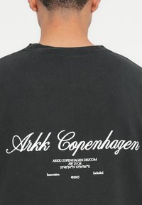 T-shirt in cotone nero con testo bianco sul retro, che include "Arkk Copenhagen" e coordinate, con colletto a girocollo e maniche corte.