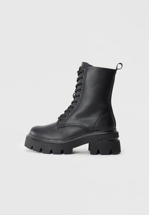 Botas de cuero negro con cordones, suela gruesa, puntera redondeada y parte superior texturizada. Presentan ojales de metal y una pequeña lengüeta en la parte trasera.
