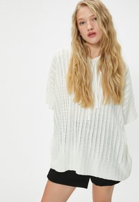 Biały, dzianinowy, oversize'owy sweter z teksturowanym wzorem i szerokim dekoltem, w zestawie z czarnymi szortami. Miękka, lekka tkanina.