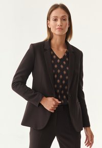 Schwarzer maßgeschneiderter Blazer mit scharfen Revers, Ein-Knopf-Verschluss und taillierter Passform, kombiniert mit einem gemusterten V-Ausschnitt-Oberteil in erdigen Farbtönen.