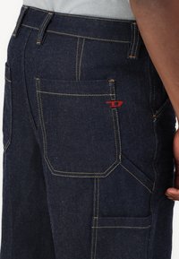Mørke denimbukser med kontrasterende gul søm, med en sidelomme og et lille rødt logodetalje nær baklommen.