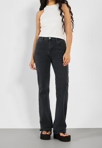 Schwarze, ausgestellte Jeans mit fünf Taschen, Knopfverschluss und Gürtelschlaufen. Getragen mit einem weißen, gerippten Trägershirt und Plateausandalen.