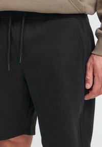 Shorts negri din bumbac, cu talie ajustabilă prin șnur, având o textură fină și buzunare laterale, asortați cu un top maro cu mânecă lungă.