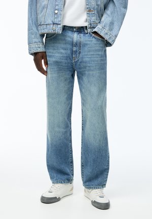 Hombre con chaqueta vaquera azul claro y vaqueros, con zapatillas blancas, de pie con una mano en el bolsillo sobre un fondo simple.