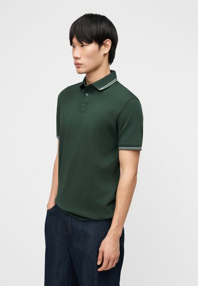 Jeune homme portant un polo vert foncé avec un bord blanc au col et aux manches, debout les mains dans les poches d'un jean foncé sur un fond uni.