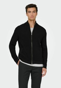 Crni rebrasti zip-up kardigan s visokim ovratnikom, prednjim džepovima i dugim rukavima, nosi se preko sive majice, u kombinaciji s tamnim hlačama.