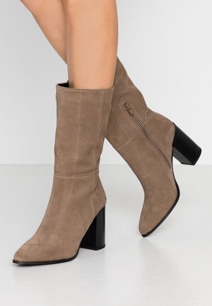 Polven korkeuteen ulottuvat taupenväriset mokkanahkasaappaat, joissa on teräväkärkinen päällinen ja teksturoitu musta blokkikorko; sivuvetoketju helpottaa pukemista.