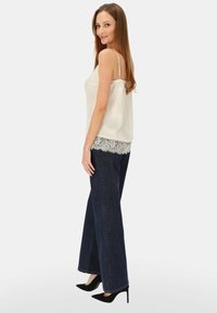 Jasnobeżowa koszulka na ramiączkach z koronkową lamówką, zestawiona z ciemnoniebieskimi spodniami o szerokich nogawkach. Modelka stoi bokiem, ma na sobie czarne szpilki z ostrym noskiem.