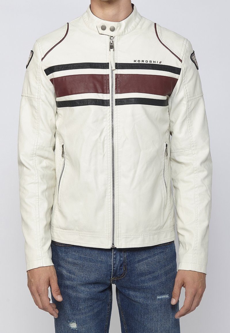 Uomo che indossa una giacca di pelle bianca con strisce orizzontali nere e marroni e chiusura lampo frontale, abbinata a jeans blu consumati.