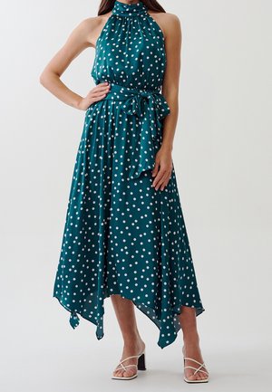 Teal halter neck midi dress with white polka dots, asymmetrical hem, tied waist sash, worn with white strappy block heel sandals.