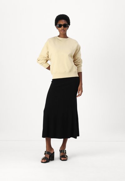 Ανοιχτό μπεζ κου sweatshirt, μαύρη φούστα maxi και μαύρες σανδάλια με τακούνι και χρυσές λεπτομέρειες. Λείες υφές και χαλαρή εφαρμογή. Καθαρό, minimal φόντο.