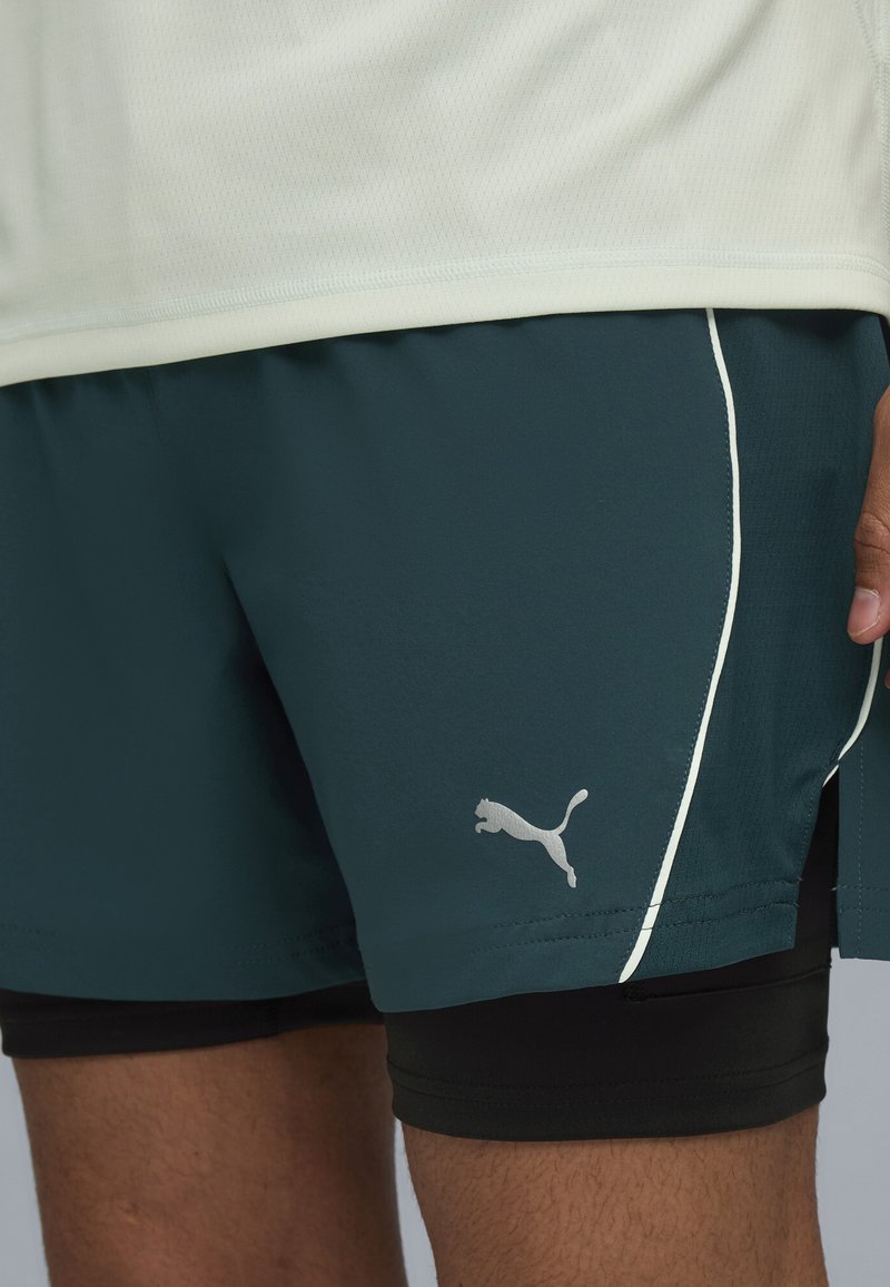Teal sportshorts met een zachte textuur, witte accenten aan de zijkanten en een grijs Puma-logo. Draag ik bovenop zwarte compressie-shorts.