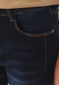 Tumedat sinised teksapüksid sujuva tekstuuriga, millel on kontrastne oranž õmblus ja esikoti detail.