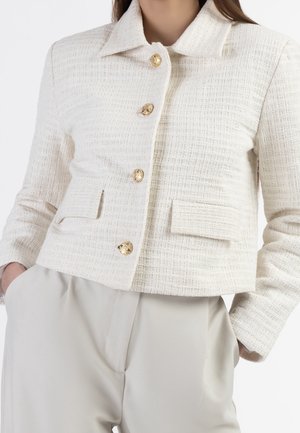 Femme portant une veste courte crème texturée avec des boutons dorés et des poches avant à rabat, associée à un pantalon beige clair.