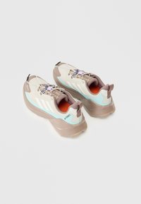 Lichte trailrunning schoenen in beige en teal, met een mesh bovenkant, een gevoerde zool en een ingewikkeld vetersysteem met hardware-accenten.