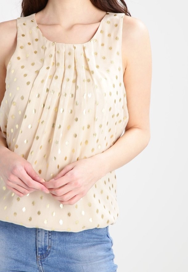 Blouse beige sans manches avec pois dorés et plis portée avec un jean bleu, mains jointes à la taille.