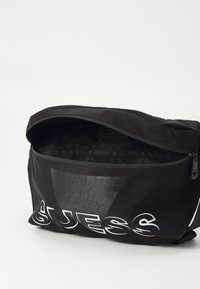 Fekete szövet táska cipzáras zárással, fényes, háromszög alakú díszítéssel és feltűnő fehér "GUESS" logóval az elején. A bélés mintás.