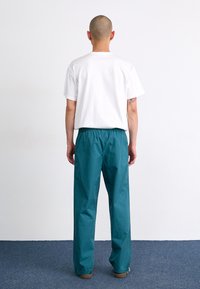 T-shirt blanc à manches courtes associé à un pantalon léger teal avec une taille élastique, présenté de dos devant un fond uni.