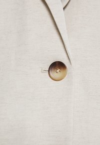 Närbild av beige tyg med en enda rund, brun graderad knapp sydd nära en slag på ett plagg.