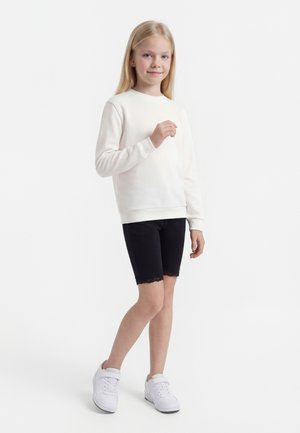 Joven niña de pie, con sudadera blanca, pantalones cortos negros con encaje y zapatillas blancas sobre un fondo blanco liso.