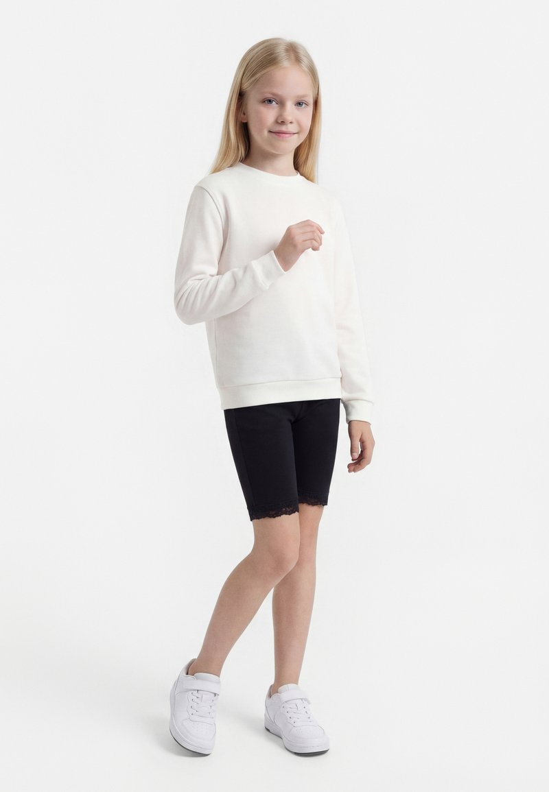 Νεαρή κοπέλα στέκεται, φοράει λευκό φούτερ, μαύρο σορτς με δαντέλα και λευκά αθλητικά παπούτσια μπροστά σε απλό λευκό φόντο.