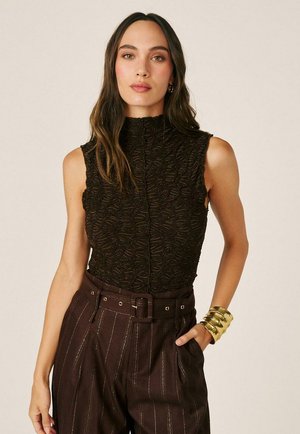 Mujer con cabello largo y oscuro, lleva una blusa negra sin mangas y con textura, pantalones altos de rayas marrones y una amplia pulsera ancha dorada.