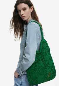 Grön virkad tote-väska med blommönster och strukturerade accenter, med hållbara handtag. Modellen bär en randig skjorta och denim.