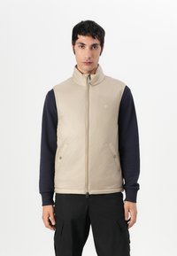 Beige isolierte Weste mit hohem Kragen und Reißverschluss, versehen mit Druckknopftaschen, kombiniert mit einem navyblauen Langarmshirt. Flaches Design, glatte Textur.