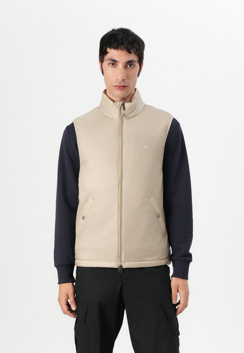 Beige isolierte Weste mit hohem Kragen und Reißverschluss, versehen mit Druckknopftaschen, kombiniert mit einem navyblauen Langarmshirt. Flaches Design, glatte Textur.