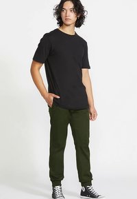 T-shirt noir à manches courtes associé à un pantalon vert ; coupe décontractée. Tissu vert foncé avec une texture lisse ; design simple. Baskets noires.