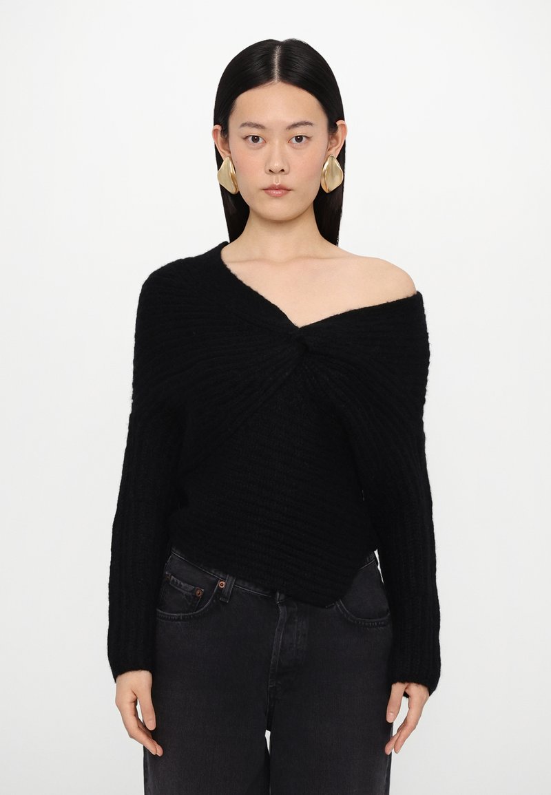 Black off-shoulder sweater with a twisted front design, ribbed texture, long sleeves, worn over dark jeans. Model displays earrings.