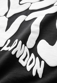Čierna tkanina s výraznými bielymi abstraktnými tvarmi a čiastočne viditeľným štylizovaným textom s nápisom "LONDON" v malých a veľkých písmenách.