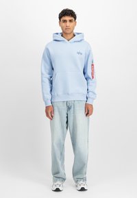 Világoskék kapucnis pulóver elülső zsebbe, bordás mandzsettákkal és a mellkason nyomott logóval. Világos mosású bő farmernadrággal és tornacipővel párosítva.