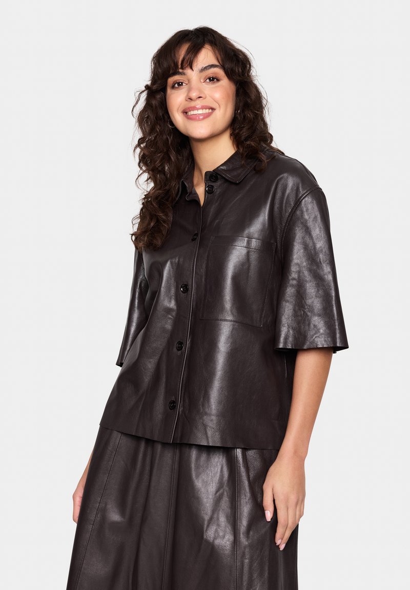 Brown leather short-sleeve shirt with a button front, pocket detail, and smooth texture. Paired with a matching leather skirt.